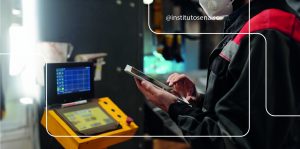 Trabalhador da indústria utilizando um tablet e uma interface digital em um ambiente de manufatura. O trabalhador veste uniforme e máscara de proteção, com um painel de controle e uma tela digital ao fundo, representando a aplicação de tecnologias da Indústria 4.0 para pequenas e médias indústrias. O texto '@institutosenaisc' está visível na parte superior da imagem.